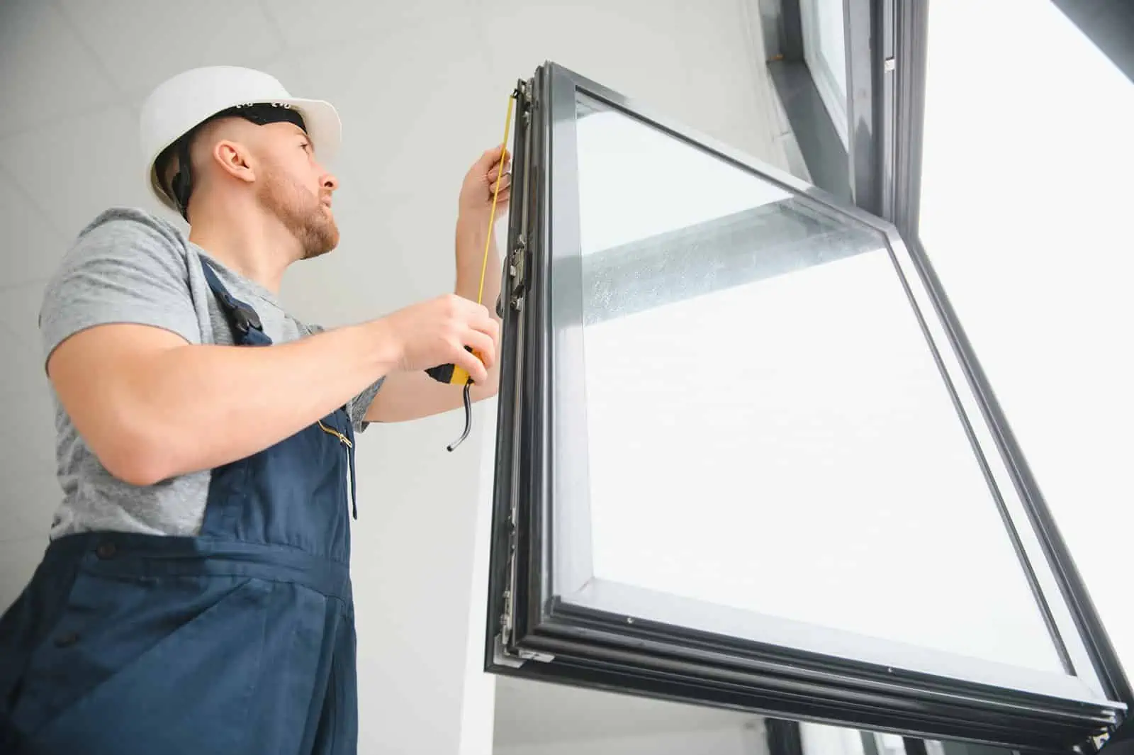 A man providing services working on a window while wearing overalls and a white hat.